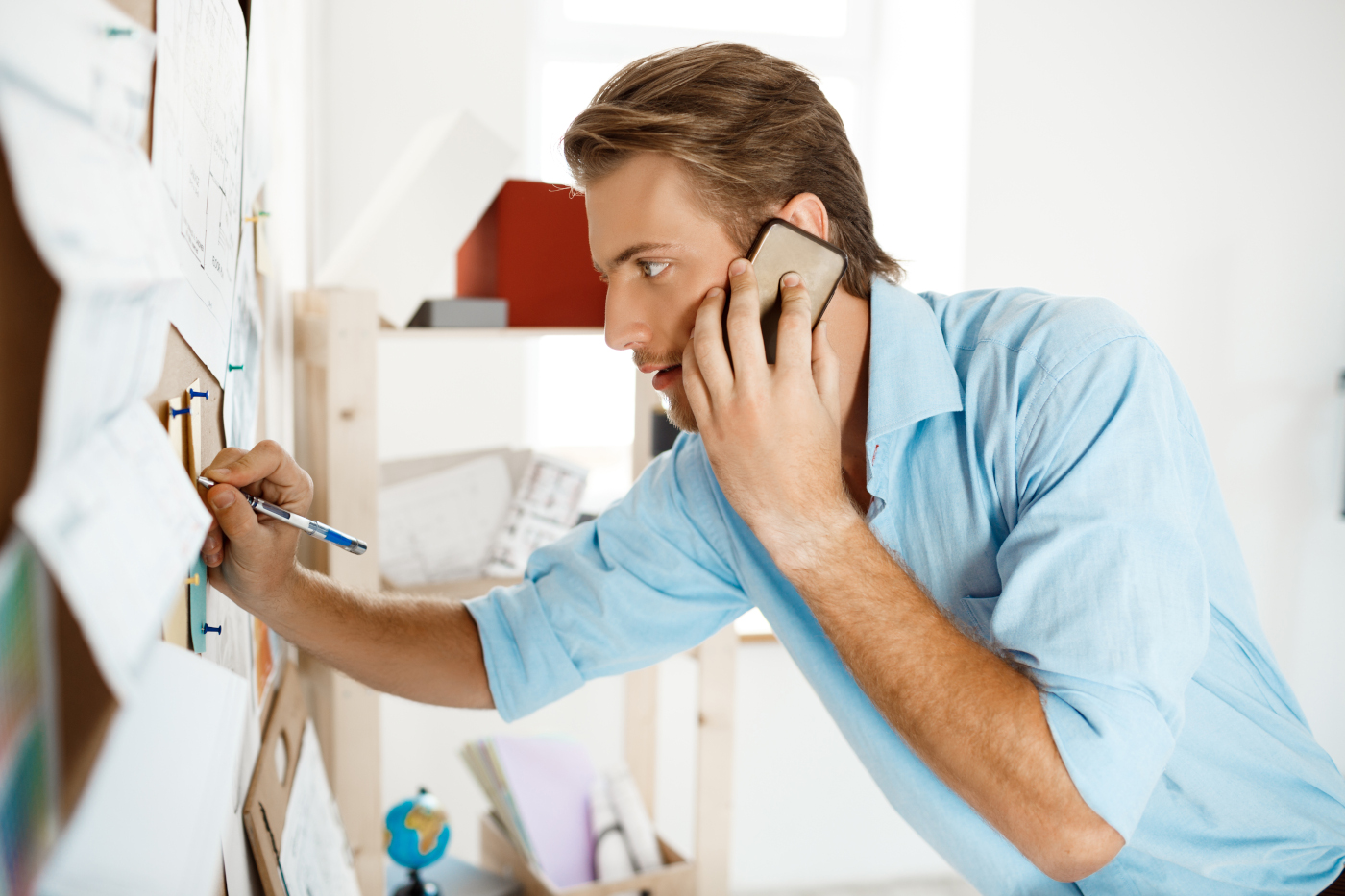 Young handsome businessman writing at the paper pinned to corkboard, talking on  phone. Modern office background.